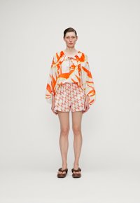 Ľahká, priesvitná blúzka s abstraktným oranžovo-bielym vzorom, skombinovaná s nohavicami so zodpovedajúcim vzorom. S outfitom ladia hnedé sandále.