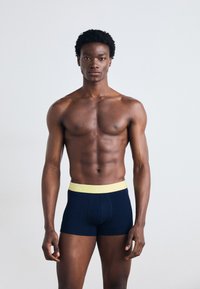 Boxers navy avec une ceinture jaune Lacoste. Tissu en coton doux, design ajusté, longueur mi-cuisse. Le modèle se tient debout, sans haut.