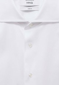 Camiseta polo blanca de corte slim fit con una textura suave, presenta un collar clásico y una tapeta de tres botones. Etiqueta de la marca visible en el interior del cuello.
