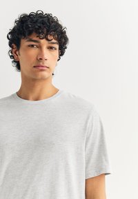 Camiseta gris claro de cuello redondo con mangas cortas, hecha de tejido suave. Presenta un ajuste relajado y una textura mínima. Sin patrones visibles.