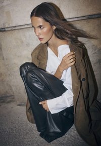 Manteau en suede marron sur une chemise blanche, associé à un pantalon en cuir texturé noir. Le mannequin est assis en position accroupie contre un mur en béton.