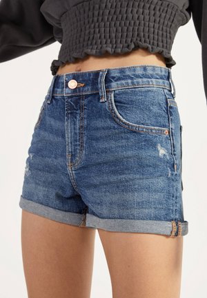 Gros plan d'une personne portant un short en jean bleu retroussé et un crop top noir texturé, montrant le ventre et le haut des jambes.