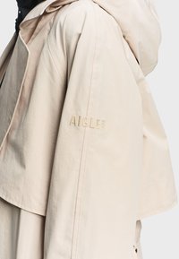 Μπεζ μπουφάν με κουκούλα, με κέντημα "AIGLE 1853" στο πάνω μέρος του μανικιού, φορεμένο πάνω από μαύρο ένδυμα.