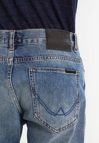 Blue denim jeans with a faded finish, featuring a leather label at the waistband and a distinct curved pocket stitching detail.