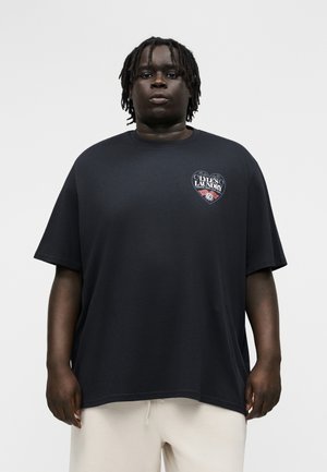 Homme noir aux dreadlocks courtes portant un T-shirt noir ample avec un petit logo "Lyle's Laundry" sur la poitrine et un pantalon de couleur claire.