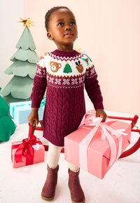 Un enfant portant une robe-pull maroon en tricot avec des motifs festifs, tenant une boîte cadeau rose. En arrière-plan, des arbres pastel et des cadeaux emballés.
