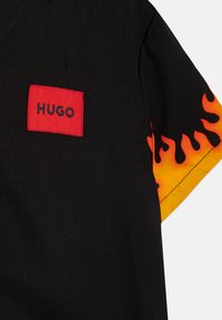 Juodas trumpomis rankovėmis marškiniai su ugnies raštu geltonomis ir oranžinėmis spalvomis ant rankovių. Raudonas stačiakampis etiketas su "HUGO" logotipu priekyje.