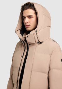 Chaqueta de plumas beige con gran capucha, con cremallera negra y cierres de botón, textura acolchada y un logotipo de acento en la manga.