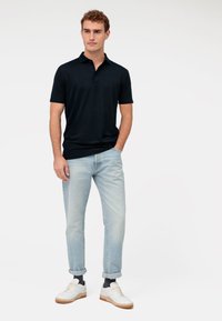 Dunkelblaues Polohemd mit Kragen, kombiniert mit hellblauen hochgekrempelten Jeans und weißen Sneakers mit braunen Sohlen. Das Outfit ist lässig und figurbetont.