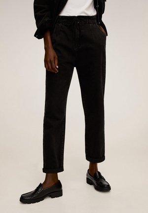 Person iført sorte bukser med manchet, sorte loafers, hvid top og sort jakke med den ene hånd i lommen mod en ensfarvet baggrund.