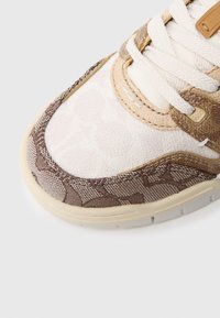 Brūna un krēmkrāsas sporta apavi ar teksturētu augšdaļu, ģeometrisku rakstu, plakanām šņorēm un bēša gumijas zoli.