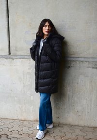 Čierna prešívaná bunda s kapucňou, so zipsom na prednej strane. Kombinovaná s modrými rozšírenými džínsami a bielymi teniskami.