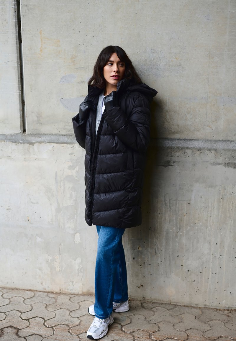 Čierna prešívaná bunda s kapucňou, so zipsom na prednej strane. Kombinovaná s modrými rozšírenými džínsami a bielymi teniskami.