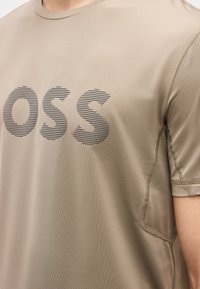 Μπεζ πουκάμισο με κοντά μανίκια και μαύρα οριζόντια ρίγες με τα γράμματα "OSS" τυπωμένα στο στήθος, που φοριέται από ένα άτομο.