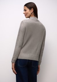 Grauer Strickpullover mit gerafftem Kragen und Bündchen, mit strukturierter Oberfläche. Kombiniert mit dunkelblauen Jeans, Ansicht von hinten.