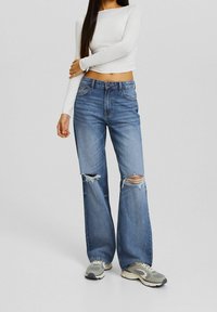 Ljust blå denimjeans med vida ben, med stora rivningar vid knäna och en urtvättad finish. Matchat med grå sneakers och en vit topp.