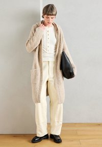 Gilet beige duveteux avec capuche, doté de deux poches avant. Porté sur un haut blanc à côtes et associé à un pantalon crème clair et des chaussures noires.