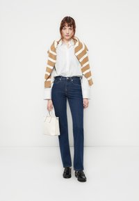 Kvinne iført hvit stripete skjorte, blå jeans, beige og hvit stripete genser over skuldrene, holder en hvit håndveske, stående mot hvit bakgrunn.