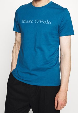 Mann trägt ein blaues Marc O’Polo T-Shirt mit kurzen Ärmeln und schwarze Hose, eine Hand in der Hosentasche und die andere entspannt an der Seite.
