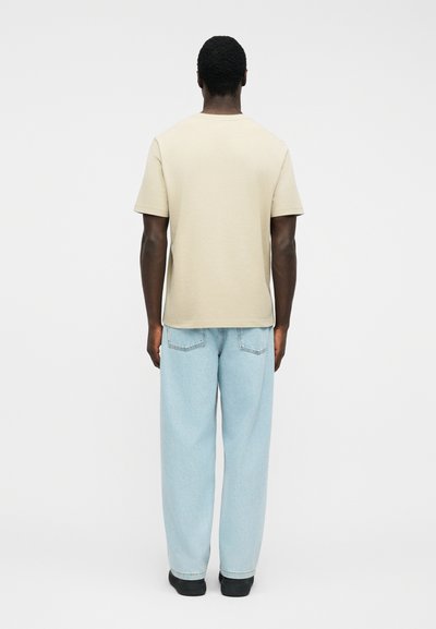 Άνδρας που φοράει μπεζ κοντομάνικη μπλούζα, ανοιχτό μπλε τζιν παντελόνι με φαρδιά μπατζάκια και μαύρα παπούτσια, στέκεται με την πλάτη προς το φακό σε απλό λευκό φόντο.