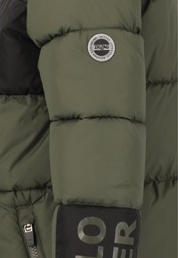 Pufjakke i olivengrøn med sorte detaljer. Har et rundt logo-patch og synlig lynlås, der viser det quiltede tekstur og isolering.