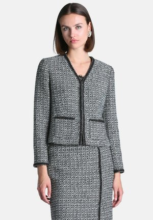 Femme portant une veste tricotée texturée noire et blanche et une jupe assortie bordée de noir, debout devant un fond clair uni.
