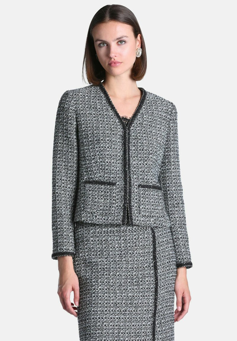 Femme portant une veste tricotée texturée noire et blanche et une jupe assortie bordée de noir, debout devant un fond clair uni.