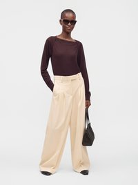 Femme portant un haut à manches longues foncé, pantalon large beige, chaussures noires, lunettes de soleil, tenant un sac à main noir, debout devant un fond blanc.