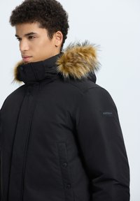 Giacca invernale nera con cappuccio foderato in pelliccia. Presenta una chiusura con zip e bottoni, tasche laterali e una patch del logo sulla manica. Tessuto testurizzato.