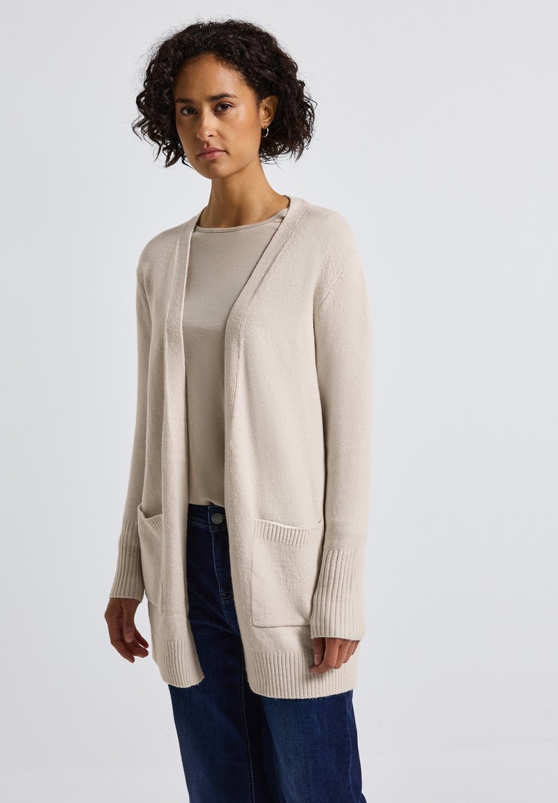 Gilet en tricot beige avec une ouverture à l'avant, des poignets côtelés et deux poches avant. Texture douce, coupe décontractée, associé à un haut léger et un jean.