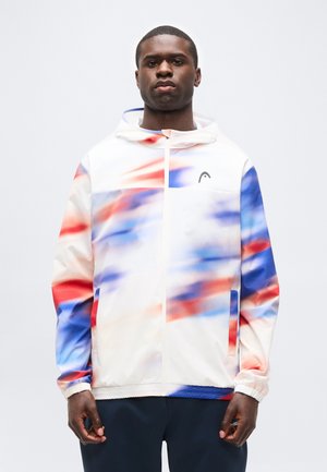 Jonge man die een witte capuchonvest met ritssluiting draagt, met rode, blauwe en oranje vage strepen, staande tegen een effen lichte achtergrond.