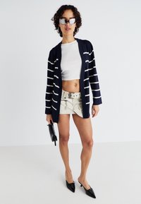 Cardigã azul-marinho com riscas brancas, top crop branco ajustado, calções bege com cinto, mala preta e saltos agulha pretos. Tecido texturizado.