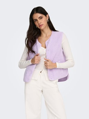 Ljus lavendelfärgad ärmlös väst med strukturerat tyg och tryckknappsstängning, lager på en vit långärmad tröja, kombinerad med vita byxor.