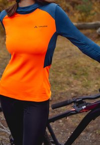 Ljusorange långärmad träningströja med blå detaljer. Slät textur, åtsittande design. Svarta cykelbyxor och mountainbike synliga.