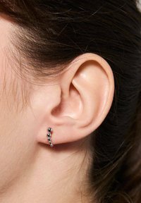 Srebrno-tonowe kolczyki z zakrzywionym wzorem, ozdobione czterema kulistymi koralikami w różnych rozmiarach, umieszczonymi na tle neutralnej karnacji skóry.