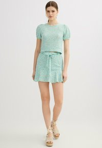 Mujer que lleva un top azul claro con flores y mangas abullonadas y una falda a juego atada, combinada con sandalias de cuña beige, de pie contra un fondo blanco.
