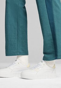 Pantaloni in tessuto a spina di pesce turchesi con una striscia laterale blu navy, abbinati a sneaker platform bianche con accenti gialli e calzini di colore chiaro.