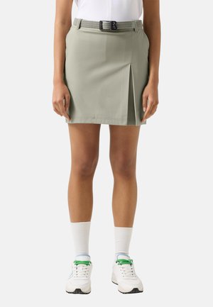 Kobieta ubrana w jasnoszarą spódnicę z rozcięciem z przodu, białe sportowe buty z zielonymi sznurowadłami oraz białe skarpetki do kostki.