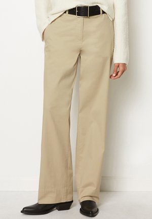 Beige broek met wijde pijpen en voorzakken, gecombineerd met een zwarte riem, witte getextureerde trui en zwarte enkellaarzen bij een persoon.