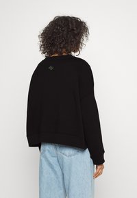 Černá oversized mikina s kulatým výstřihem a spadlou ramenou. Na horní části zad se nachází malé zelené logo. Ležérní design.