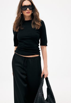 Frau mit langem braunem Haar, die schwarze Sonnenbrillen, ein schwarzes Oberteil und eine schwarze Hose trägt, hält eine große schwarze gewebte Handtasche vor einem schlichten weißen Hintergrund.