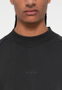 Melna sporta krekls ar ribotu kakla izgriezumu un smalku izsistētu logo. Gluda tekstūra un pieguļošs dizains, kas izceļ augšējo ķermeni.