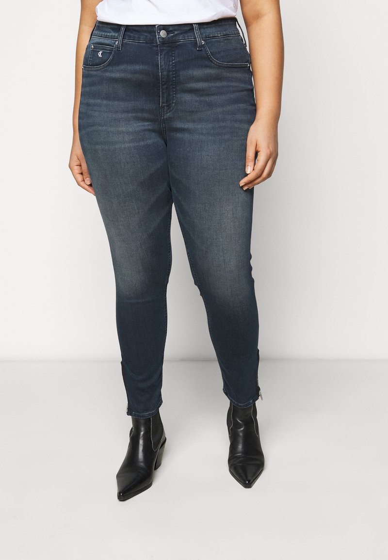 Jeans de tiro alto en azul con un corte slim, hechos de mezclilla. Presenta bolsillos frontales y cremalleras en el tobillo. Combinados con botas negras de tacón.