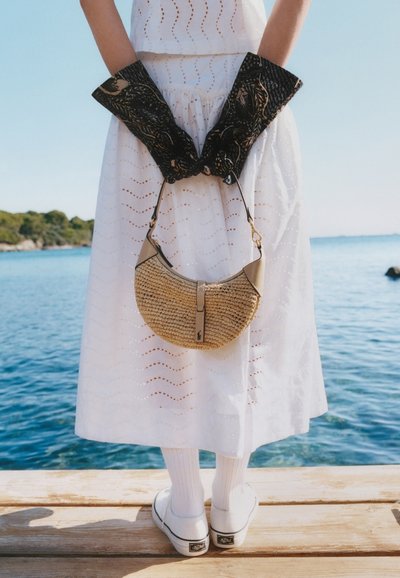 Άτομο με λευκό φόρεμα και αθλητικά παπούτσια στέκεται σε ξύλινο μόλο δίπλα στο νερό, φοράει μαύρα γάντια με σχέδια και κρατά ένα μικρό πλεκτό τσαντάκι πίσω από την πλάτη.