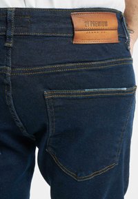 Tummansiniset farkut, joissa on näkyvää keltaista ompelenkkiä, varustettuna nahkapaikalla, jossa lukee "2Y PREMIUM JEANS CO.", sekä takataskusuunnittelulla.