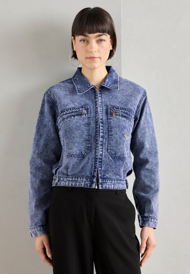 Blue denim cropped jacket with a front zipper, two chest pockets, and a light wash texture. Features a pointed collar and contrasting stitching.