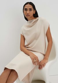 Beige Drapé-Kleid mit einem Cowl-Ausschnitt und kurzen Ärmeln, glattes Material, knielang mit asymmetrischem Saum, getragen mit einem Ring.