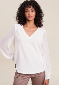 Weiße Bluse mit V-Ausschnitt, strukturiertem Stoff und langen, fließenden Ärmeln. Elastische Details an den Ärmelbündchen setzen Akzente. Getragen mit beigen Hosen.