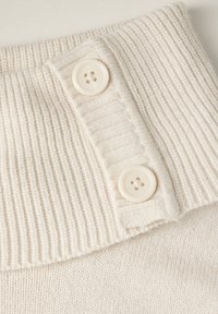 Tessuto a maglia di colore crema con colletto a coste e due bottoni circolari a quattro fori che accentuano il design. Morbido al tatto e leggero.