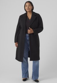 Svart lang frakk med slagkrage, løs passform og rett design, båret over en svart topp og parret med lyseblå flagrende jeans.